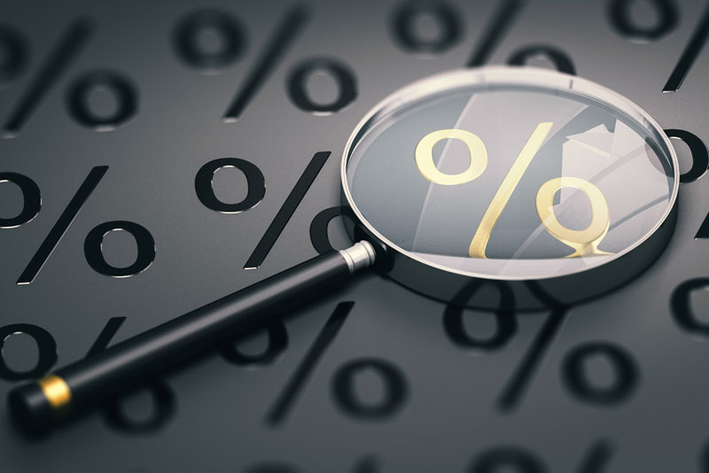 Lupe liegt auf grauer Unterlage mit Prozentzeichen darauf. Das Prozentzeichen innerhalb der Lupe ist gold.