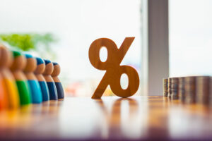 Bunte Holzfiguren, ein Prozentsymbol und gestapelte Münzen auf dem Tisch, die Zinssätze, Einkommensverteilung, Bevölkerungsfinanzierung, wirtschaftliche Ungleichheit und Finanzdemografie symbolisieren.